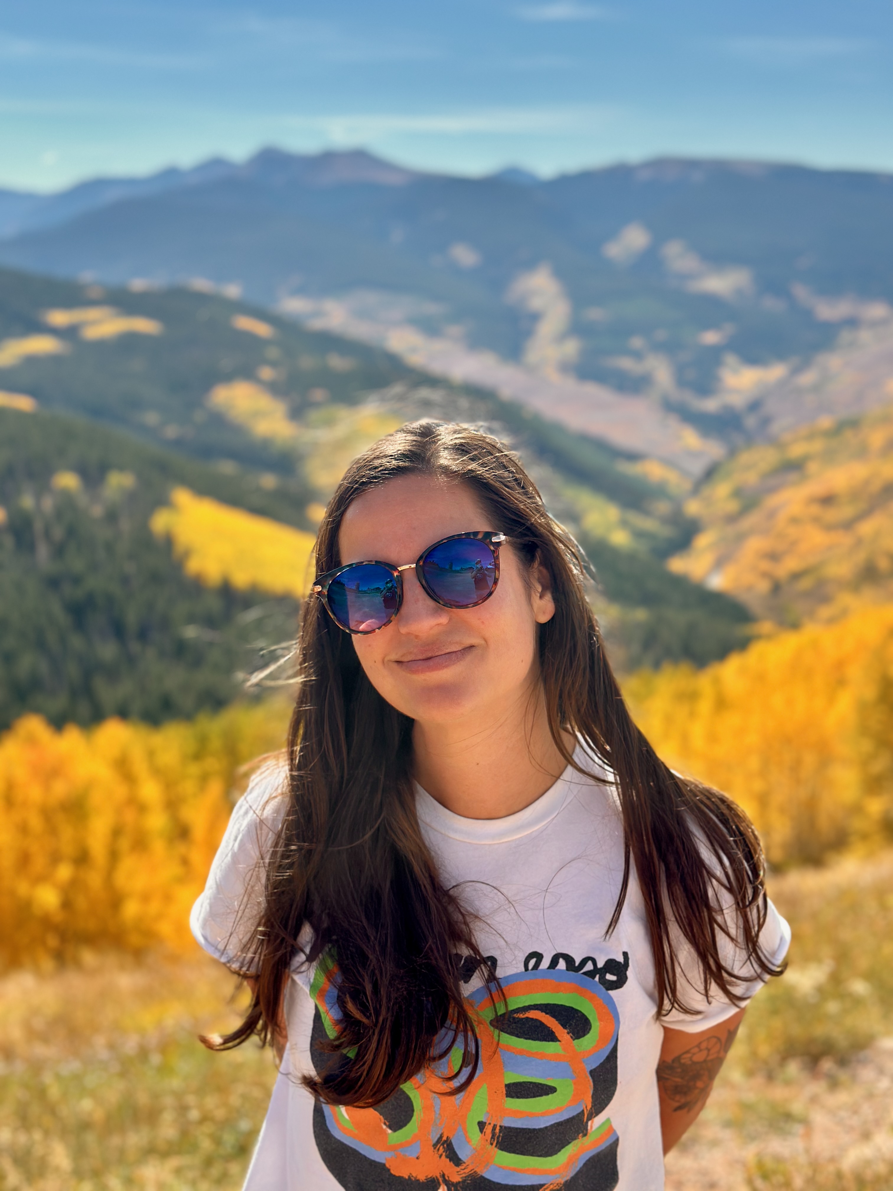 Amanda Waldrop, a birth doula, smiling on a mountain trail with autumn trees and peaks behind her