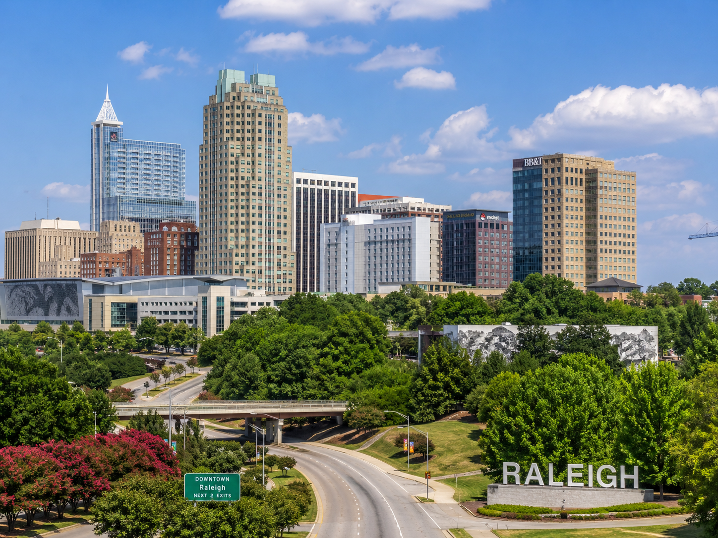 Raleigh skyline above a green park and curving road under a blue sky