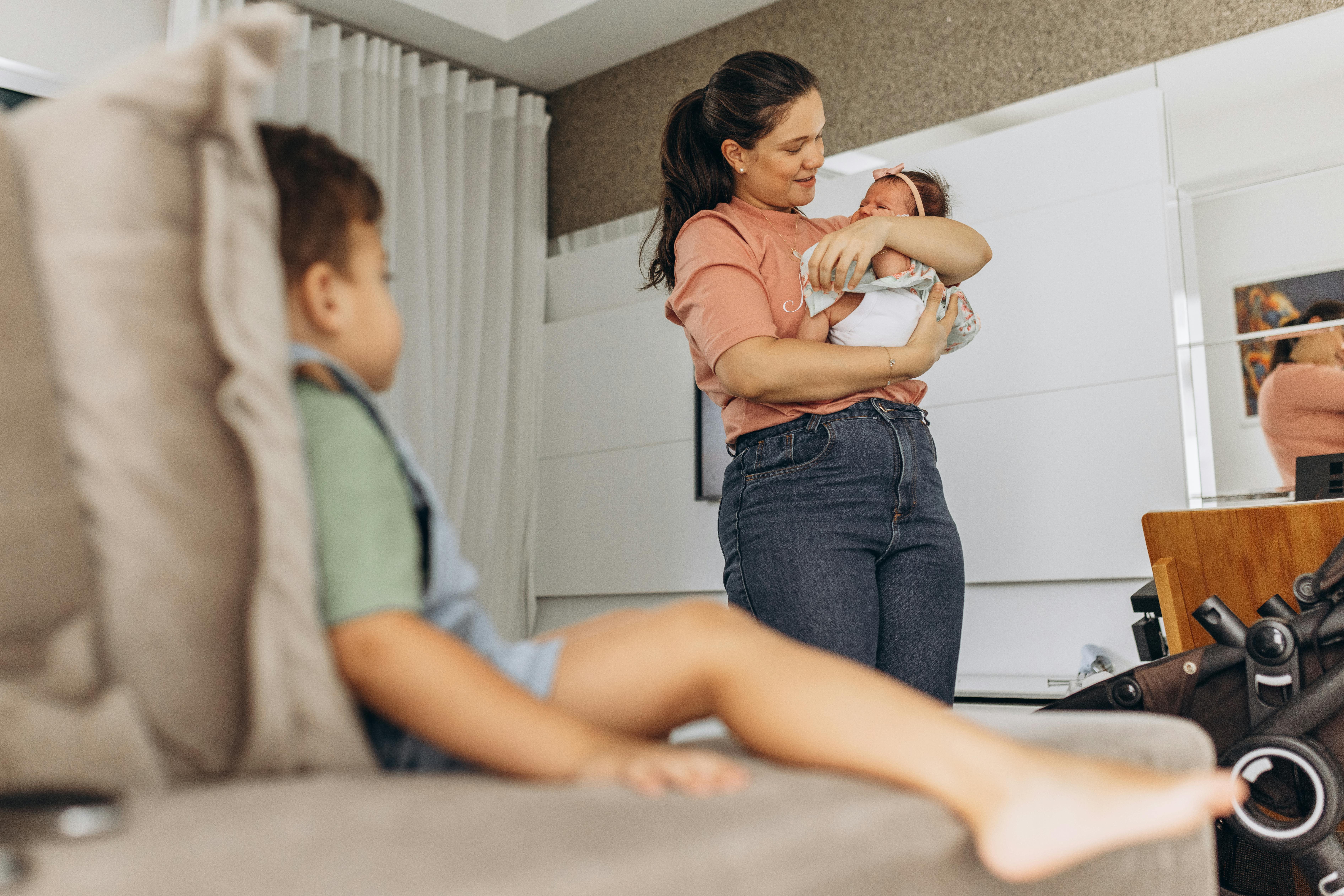 Mother holding a baby in a bright living room while a child sits on the sofa