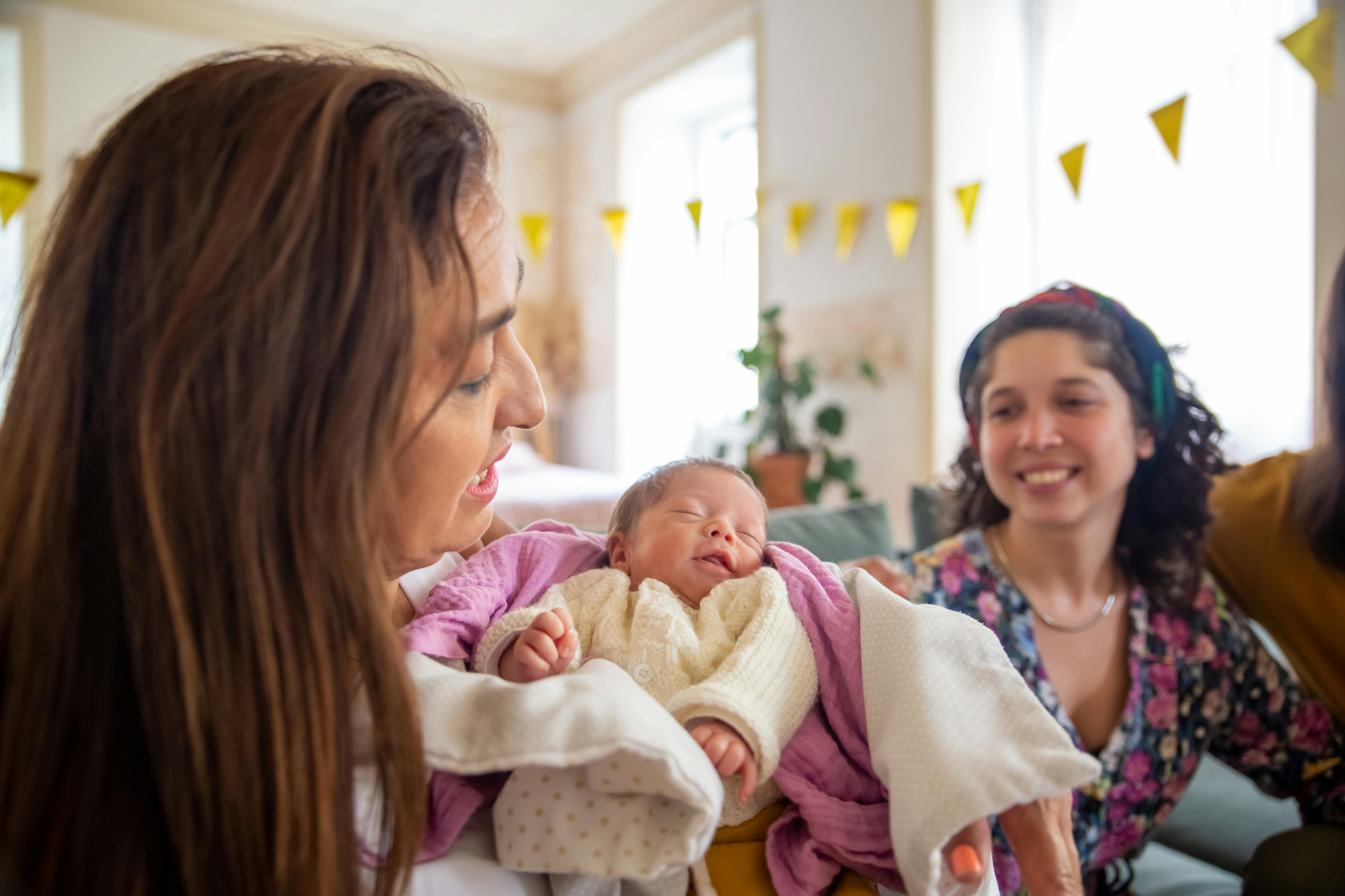 Woman holding a newborn baby while chatting with two smiling guests at a party