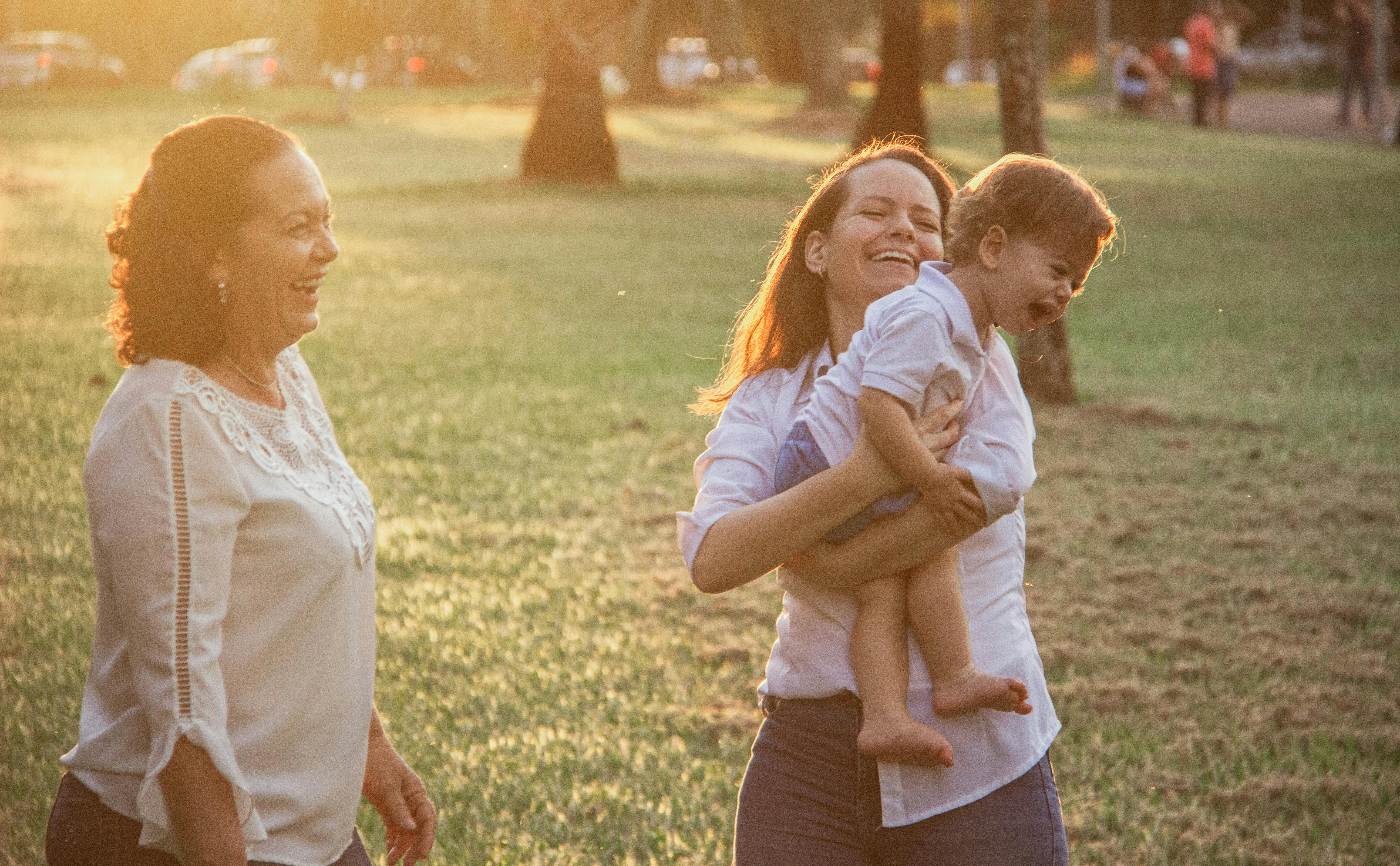 Mother carrying a young child while smiling in a sunlit park; another woman stands nearby.