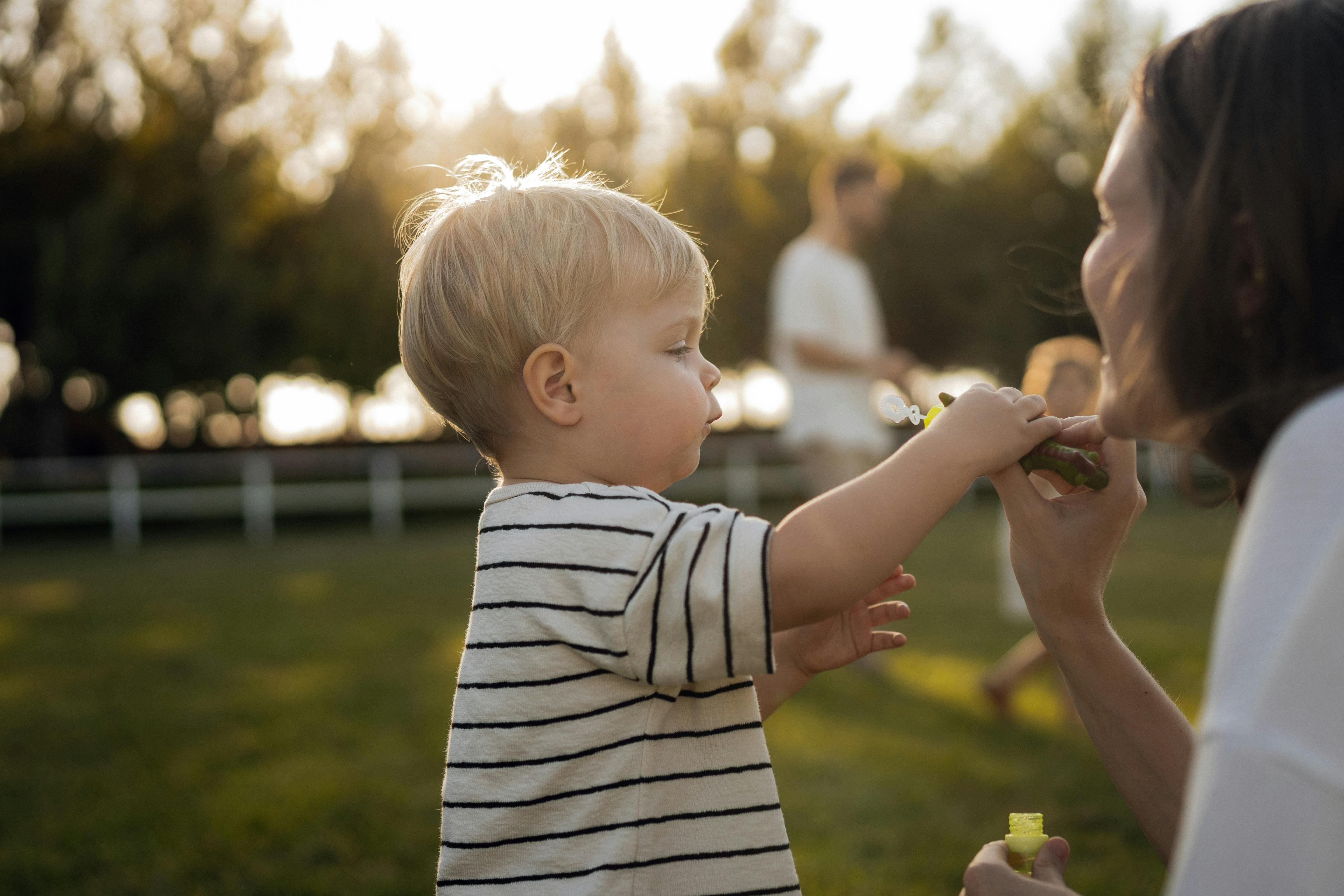Toddler reaching toward an adult&rsquo;s face in a sunlit park.
